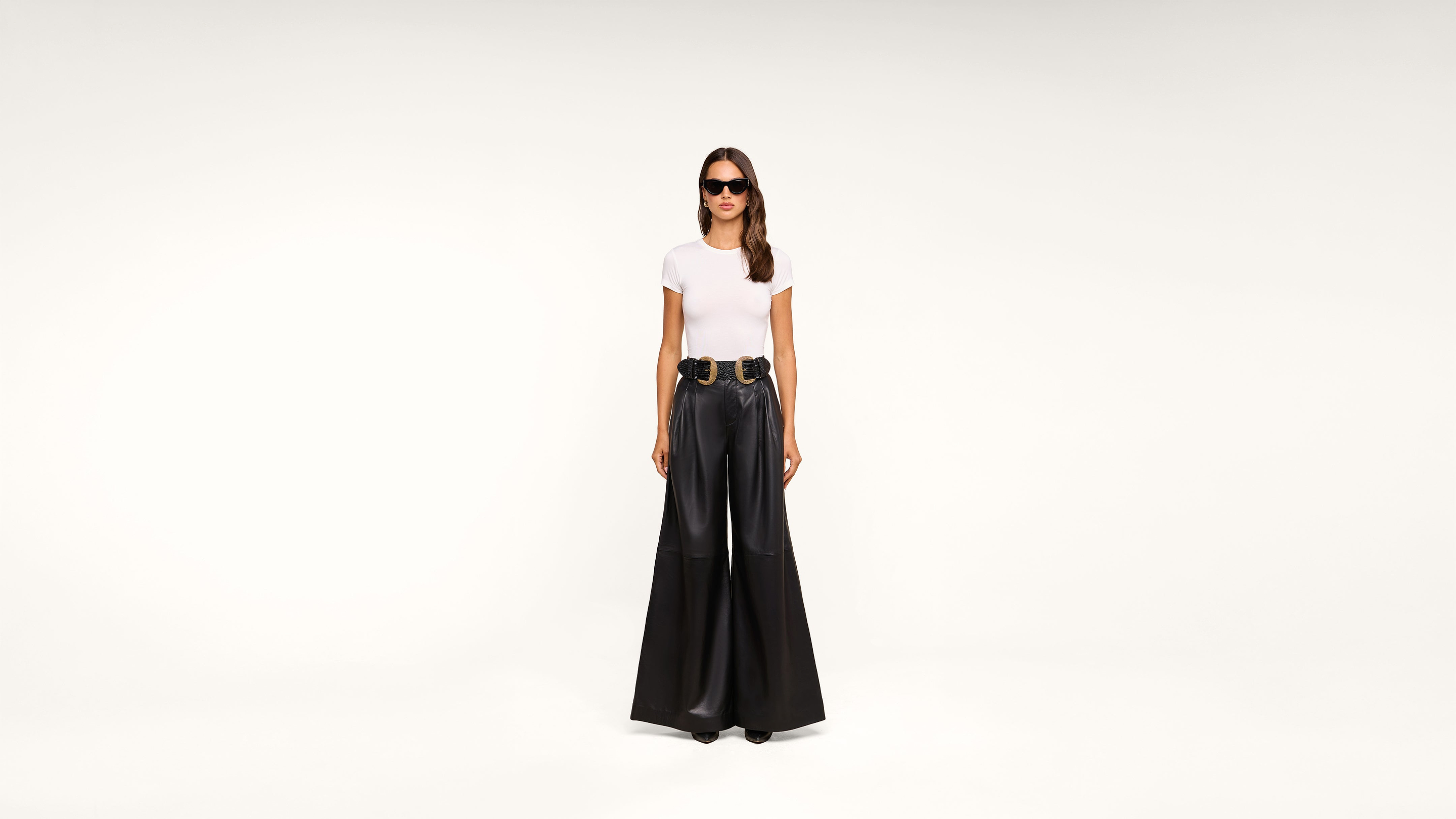 A woman wearing sunglasses, a white t-shirt, a wide black belt, and high-waisted, wide-leg black pants stands against a plain white background.