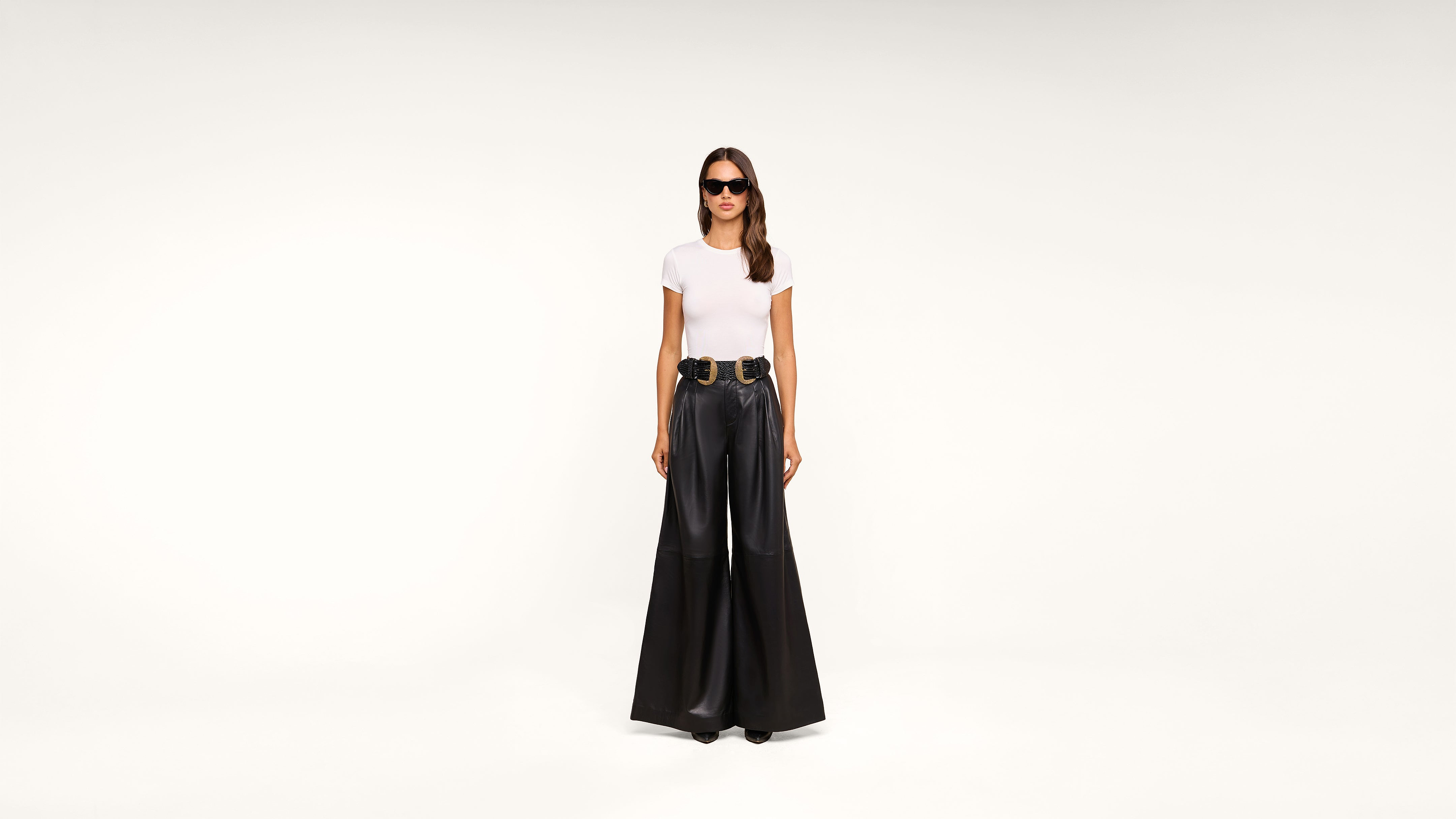 A woman wearing sunglasses, a white t-shirt, a wide black belt, and high-waisted, wide-leg black pants stands against a plain white background.