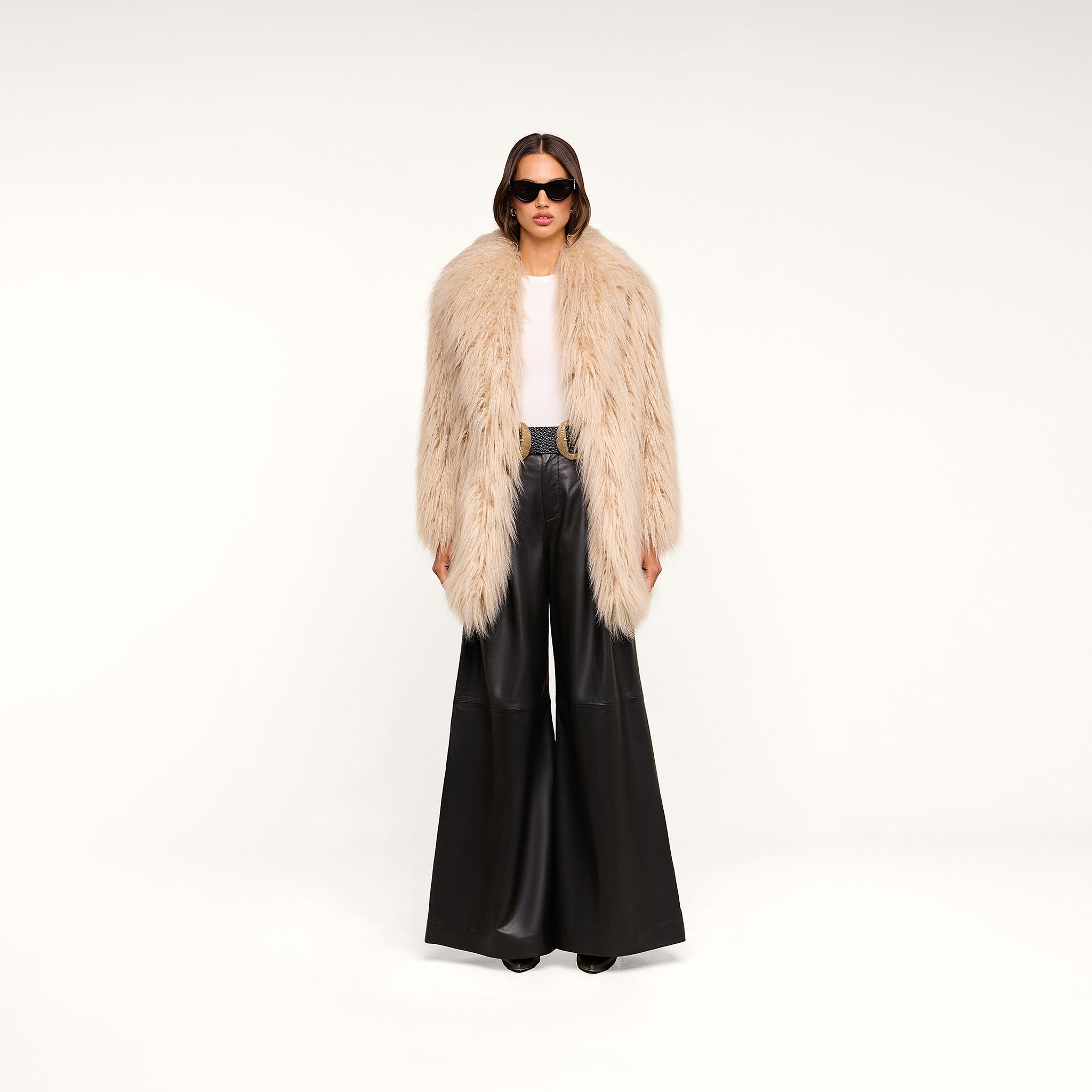 A woman stands against a plain white background, wearing a light faux fur coat, white top, black belt, sunglasses, and the Chelsy Leather Wide-Leg Pant.
