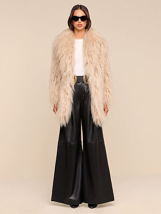 A woman stands against a light background wearing a fluffy beige faux fur coat, white top, Chelsy Leather Wide-Leg Pant, black belt with a gold buckle, black shoes, and black sunglasses.