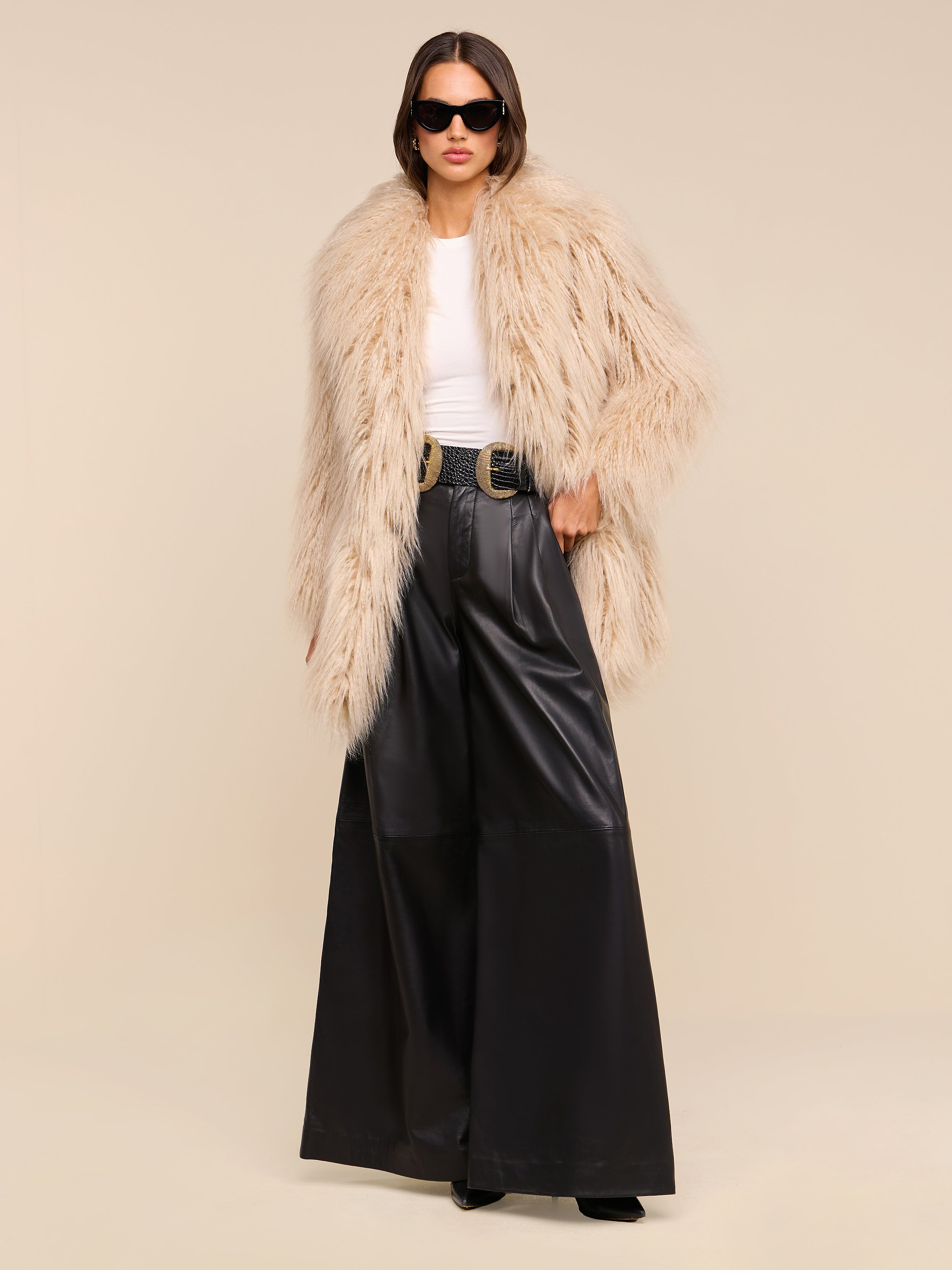 A person wearing a beige faux fur coat, white top, the Chelsy Leather Wide-Leg Pant in a high-rise fit, black boots, and sunglasses stands against a plain white background.
