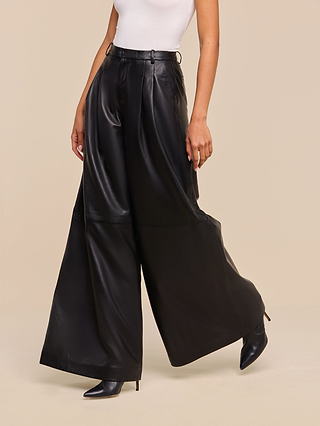 A person wearing a white sleeveless top and the Chelsy Leather Wide-Leg Pant, styled with black pointed-toe heels, poses against a beige background.