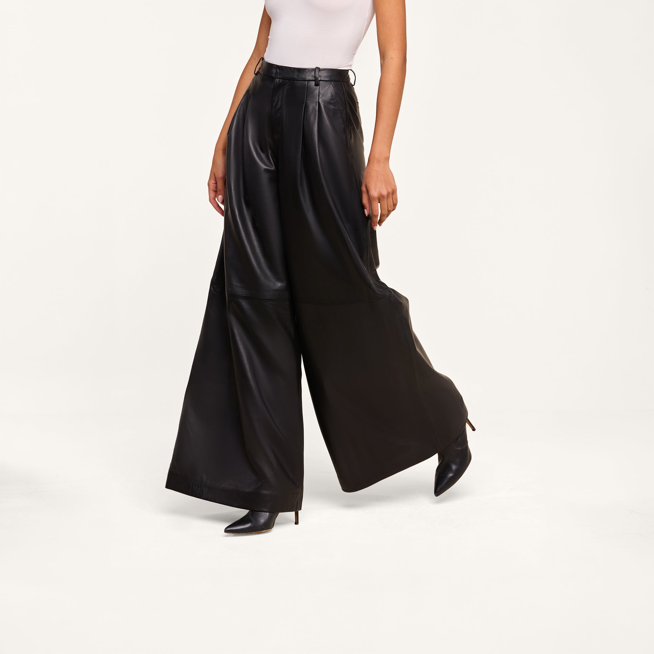 A person wearing a white sleeveless top, the Chelsy Leather Wide-Leg Pant crafted from lambskin leather, and black pointed-toe boots stands against a plain white background.