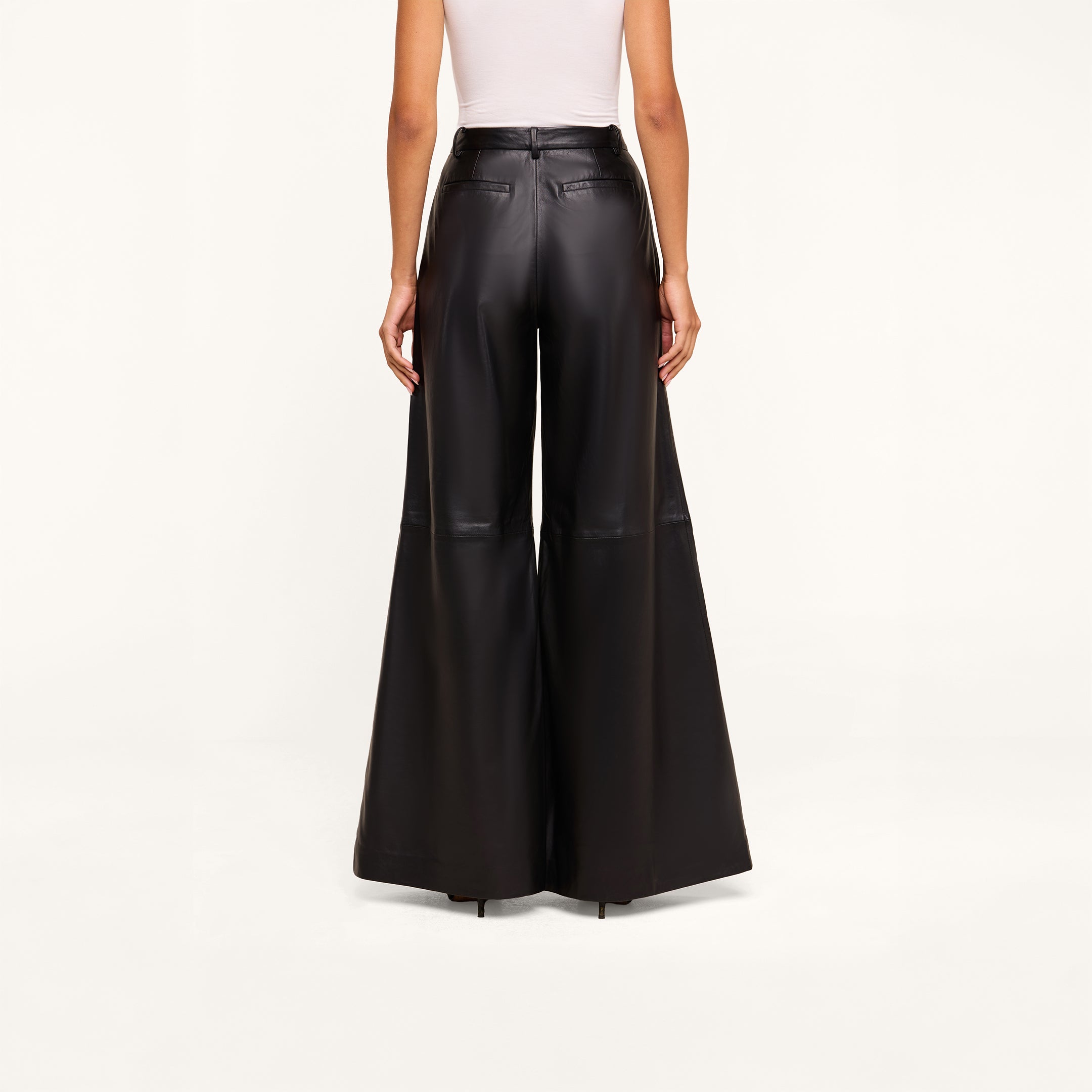 A person is wearing a white sleeveless top and high-rise, black Chelsy Leather Wide-Leg Pants made from lambskin, standing facing away against a plain light background.