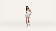 A woman wearing a white tank top, light-colored shorts, and high-heeled sandals stands with hands in pockets against a plain white background.