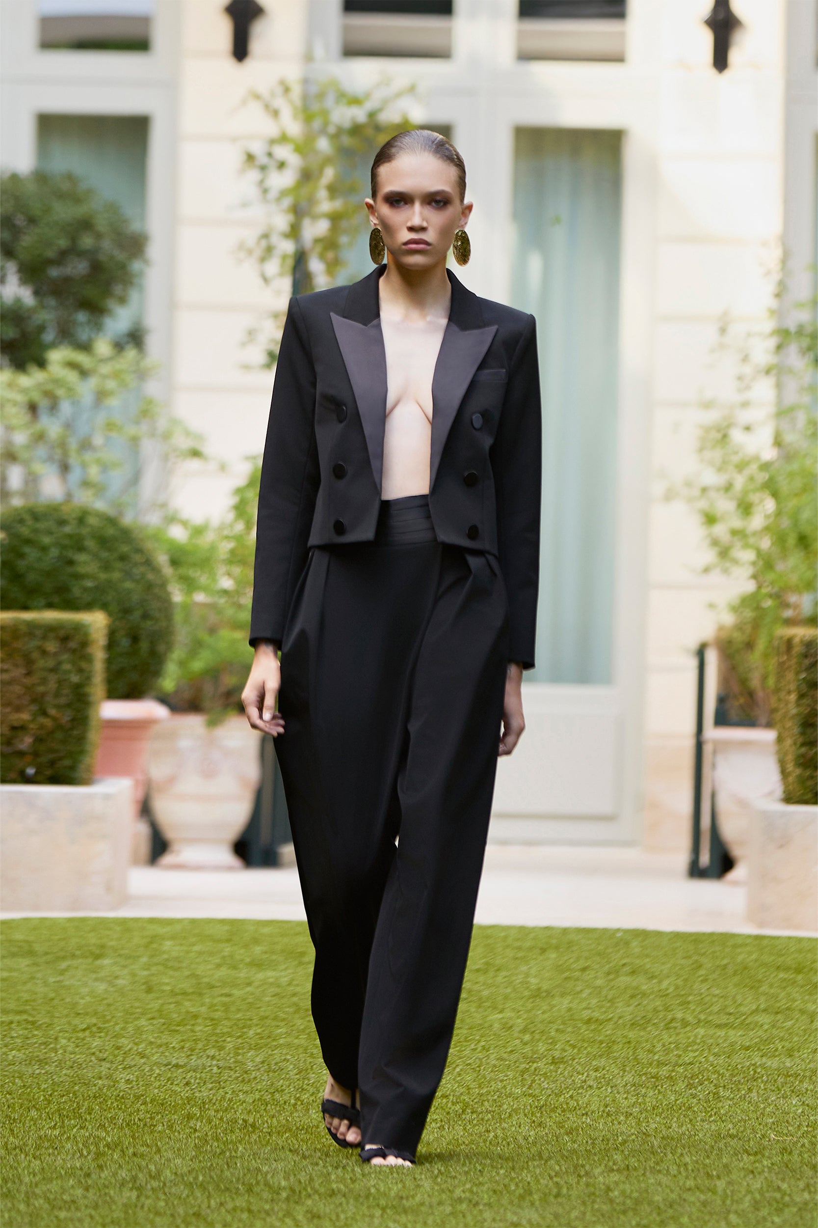 A model walks on grass in a formal outdoor setting, wearing a black double-breasted blazer with matching loose black trousers and large statement earrings.