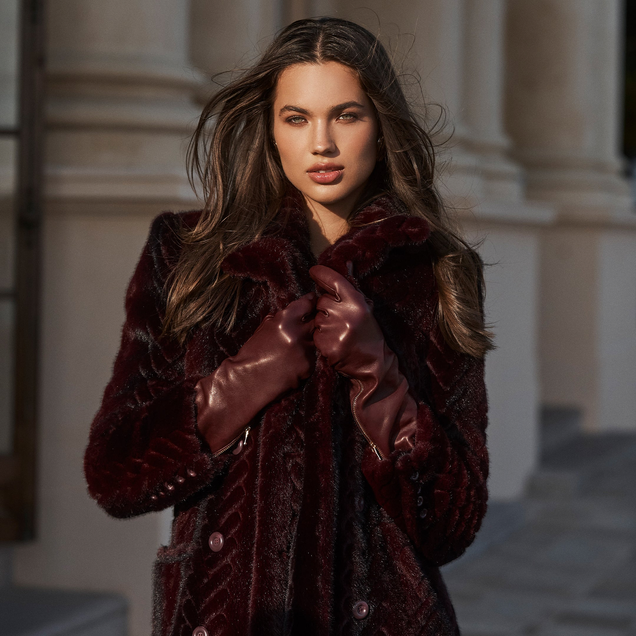 A woman with long brown hair wears a dark burgundy fur coat and matching gloves, standing outdoors in front of a light-colored building with columns, exuding elegance.