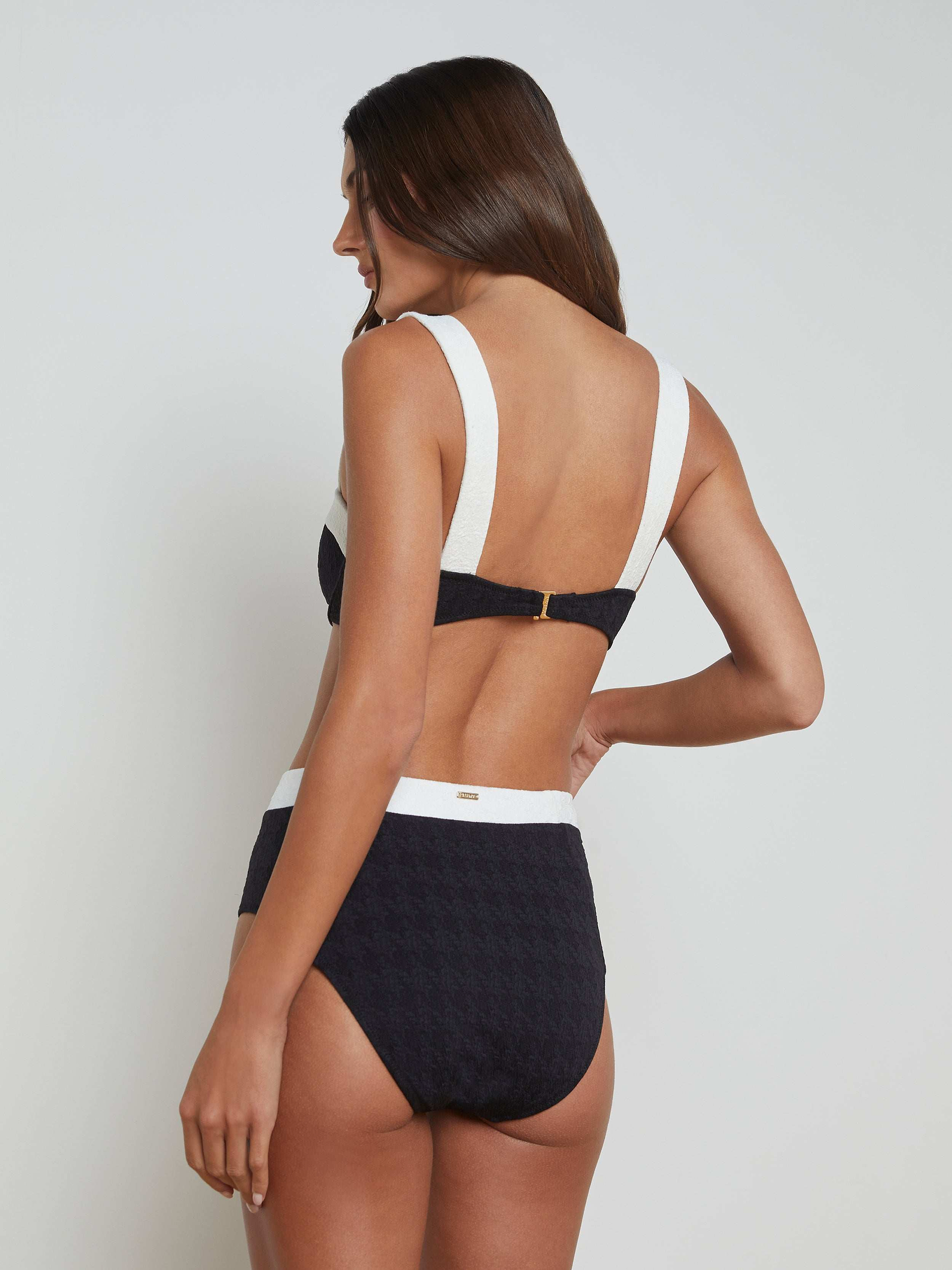 A woman with long brown hair is shown from the back against a plain light background wearing the Vanessa High-Waisted Bikini Bottom—a black and white textured bouclé piece adorned with gold buttons.