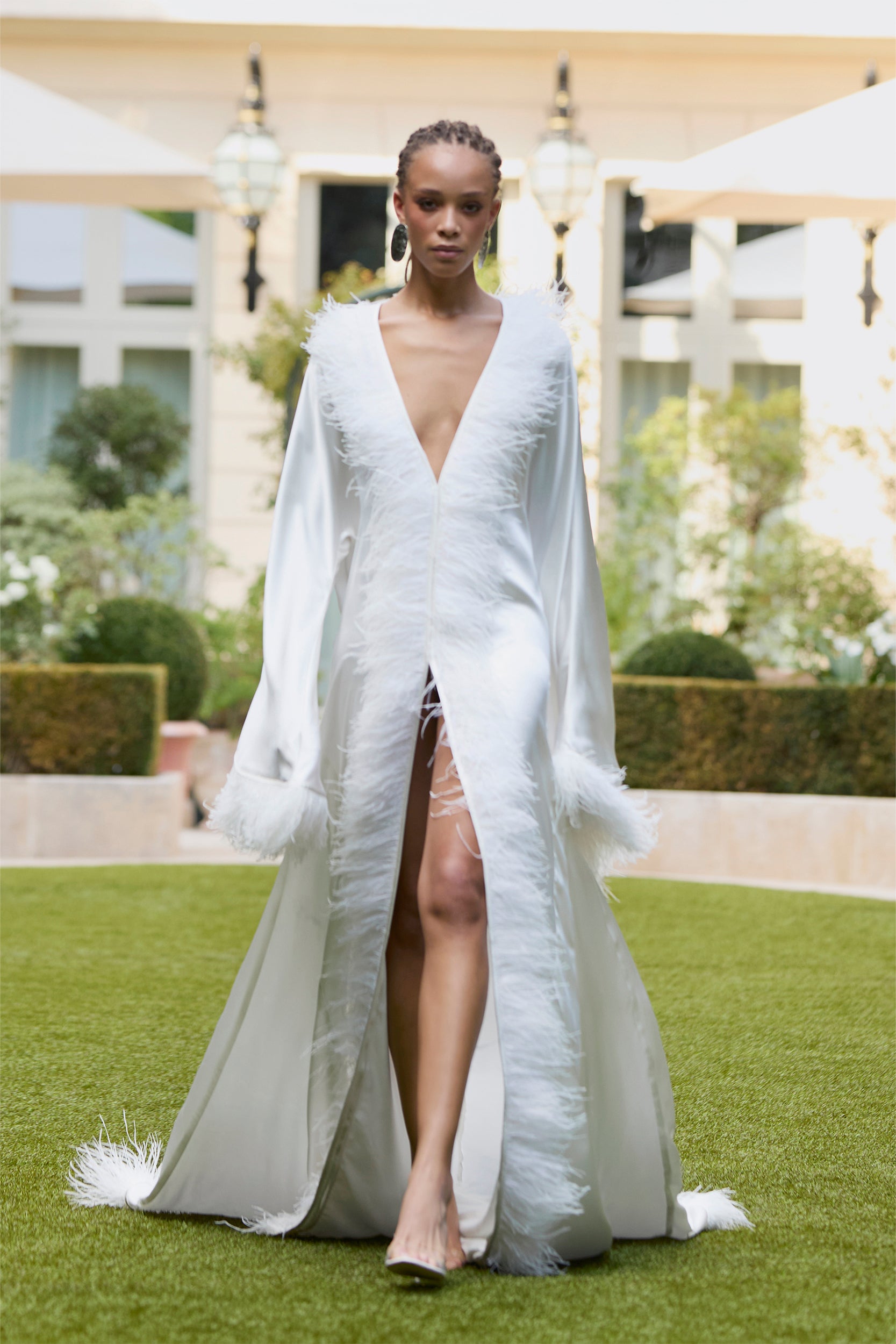 A model in a long white gown with feather trim and flowing sleeves walks on grass outdoors, framed by greenery and a building with large windows in the background.