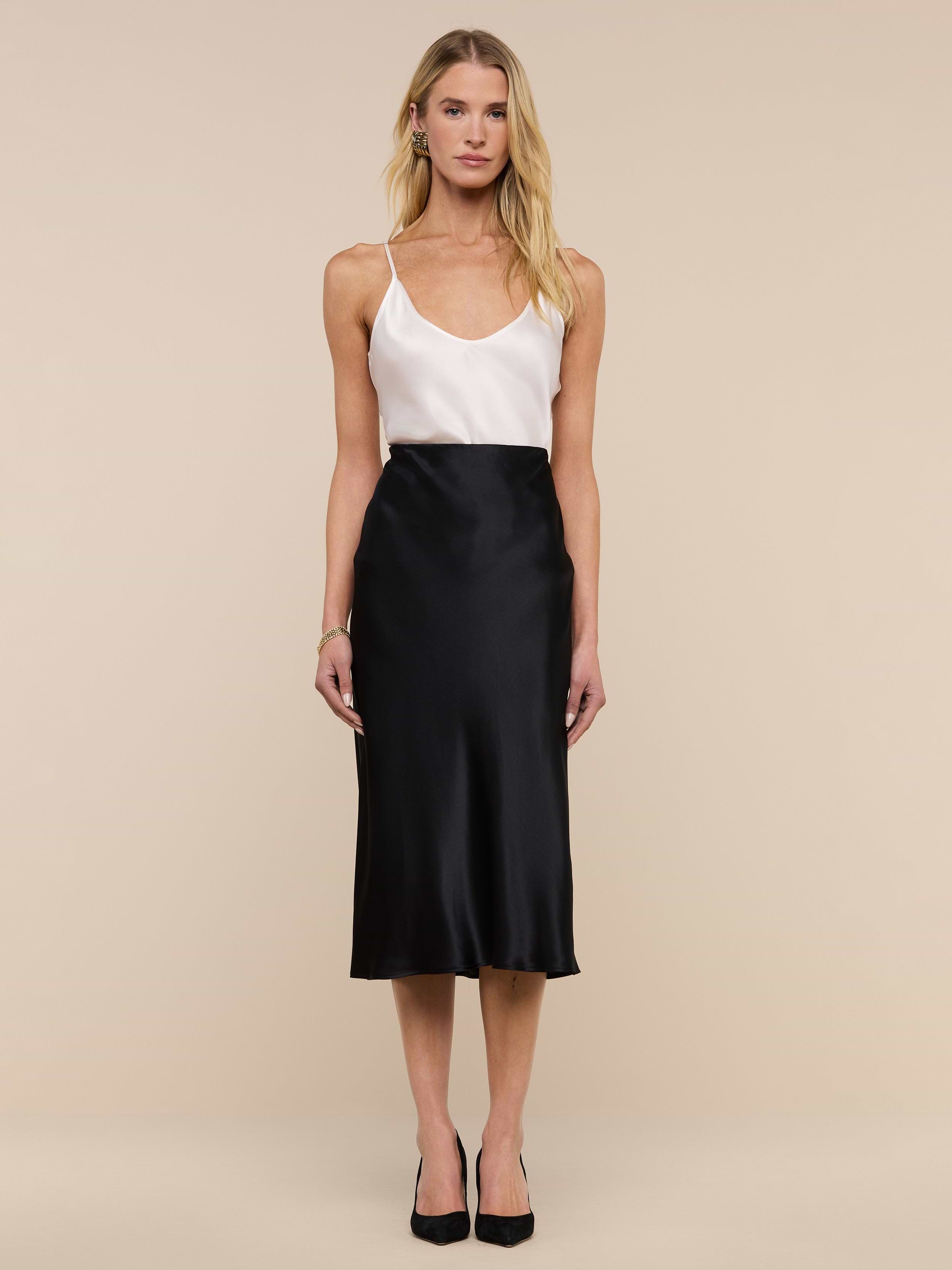 A woman stands against a plain background, wearing a white sleeveless top, the Perin Silk Midi Skirt in black, and black heels. She has long blonde hair and a neutral expression.
