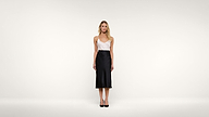 A woman stands in a minimal studio wearing a white sleeveless top, a black midi skirt, and black heels, facing forward against a plain white background.