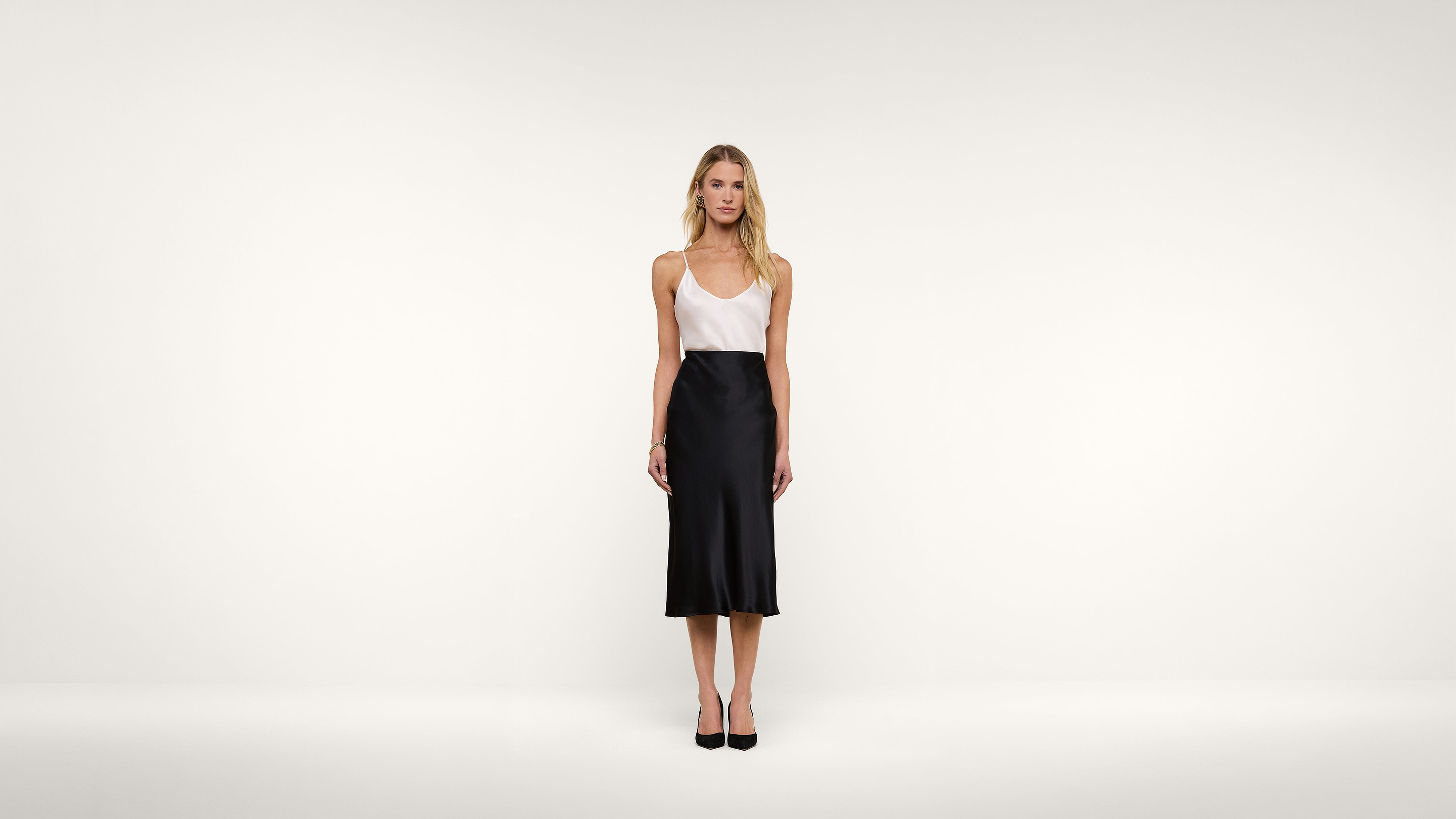 A woman stands in a minimal studio wearing a white sleeveless top, a black midi skirt, and black heels, facing forward against a plain white background.