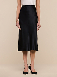 A woman wearing the Perin Silk Midi Skirt in black, paired with a white sleeveless top and black high heels, stands against a plain white background.