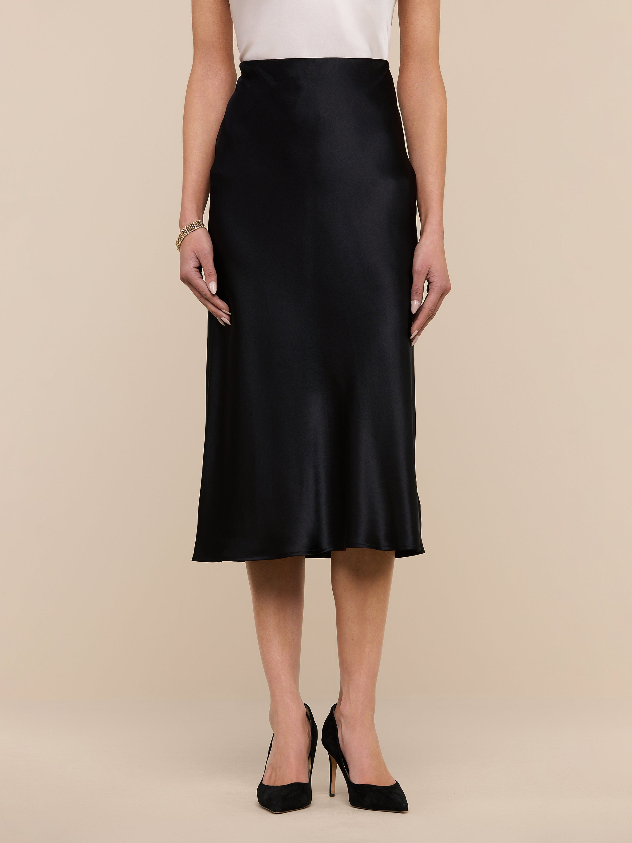 A woman wearing the Perin Silk Midi Skirt in black, paired with a white sleeveless top and black high heels, stands against a plain white background.