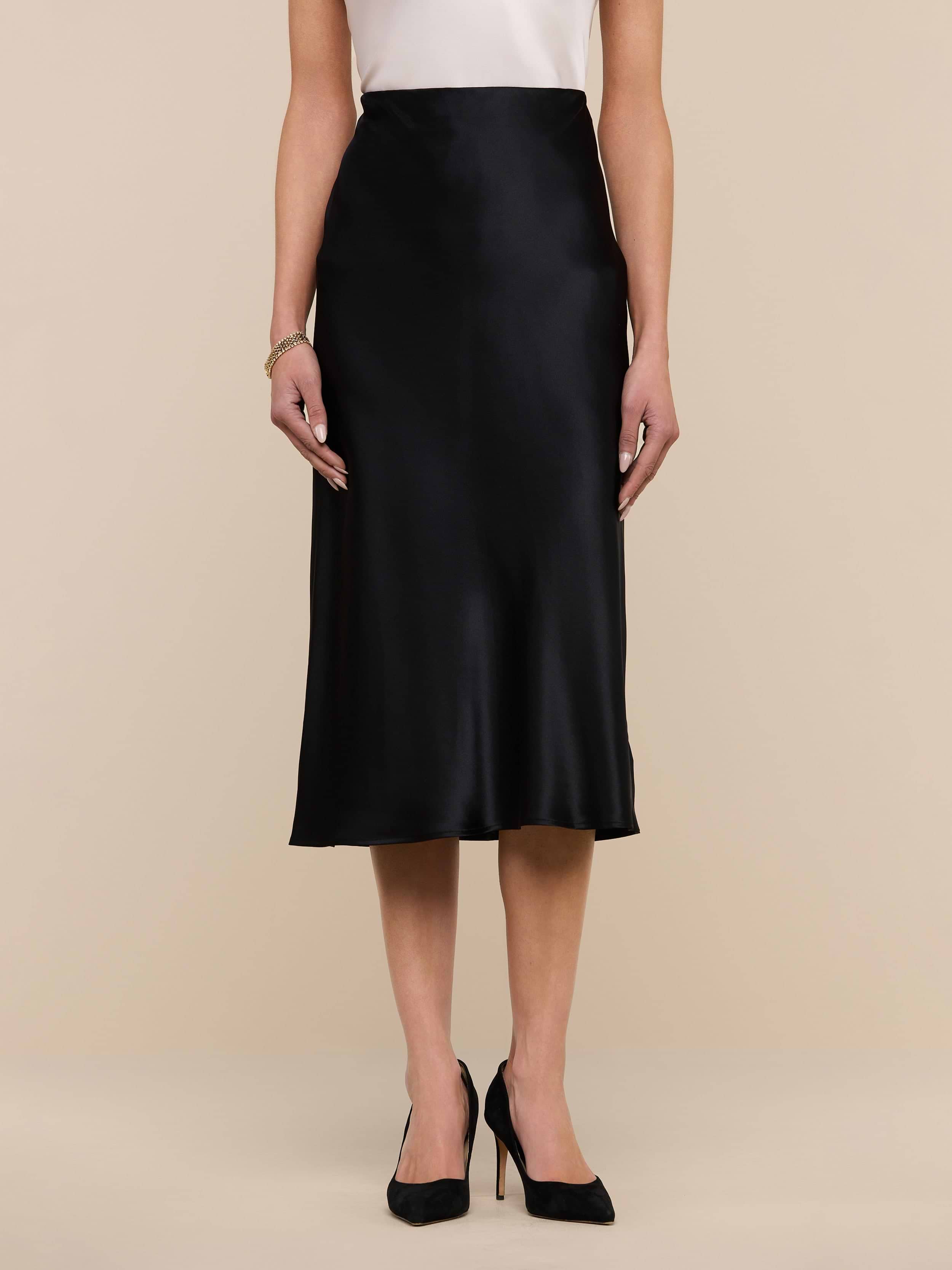 A woman wearing the Perin Silk Midi Skirt in black, paired with a white sleeveless top and black high heels, stands against a plain white background.