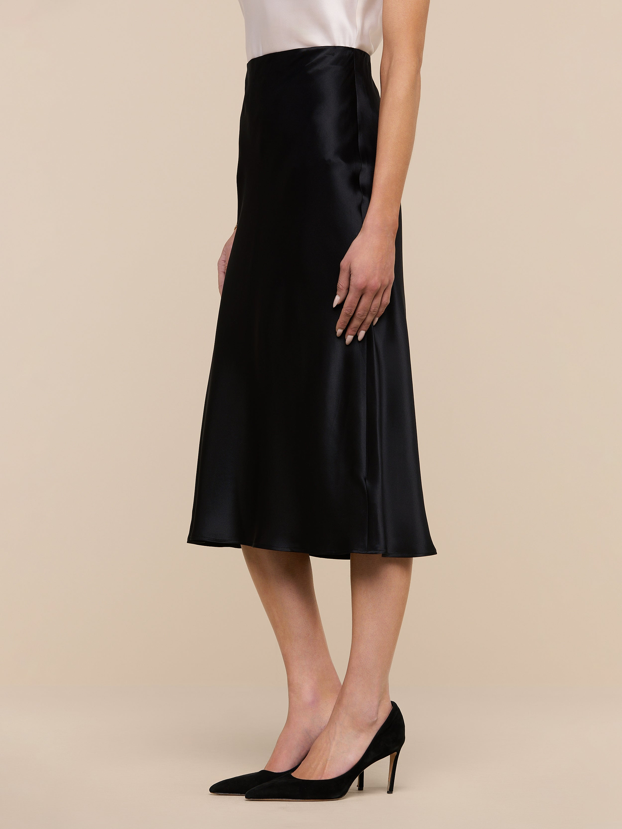 A woman wearing the Perin Silk Midi Skirt in black, styled with a white top and black high heels, standing against a plain white background.