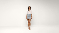 A woman stands in a minimalist white studio, wearing a light pink blouse, a blue denim mini skirt, and white high-heeled sandals. She has long brown hair and a relaxed, confident pose.