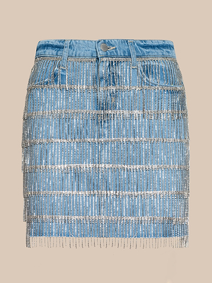 The Jolene Embellished Mini Skirt is a blue denim mini skirt decorated with rows of crystal fringe and rhinestones, plus belt loops, front pockets, and a button closure. Displayed on a beige background.