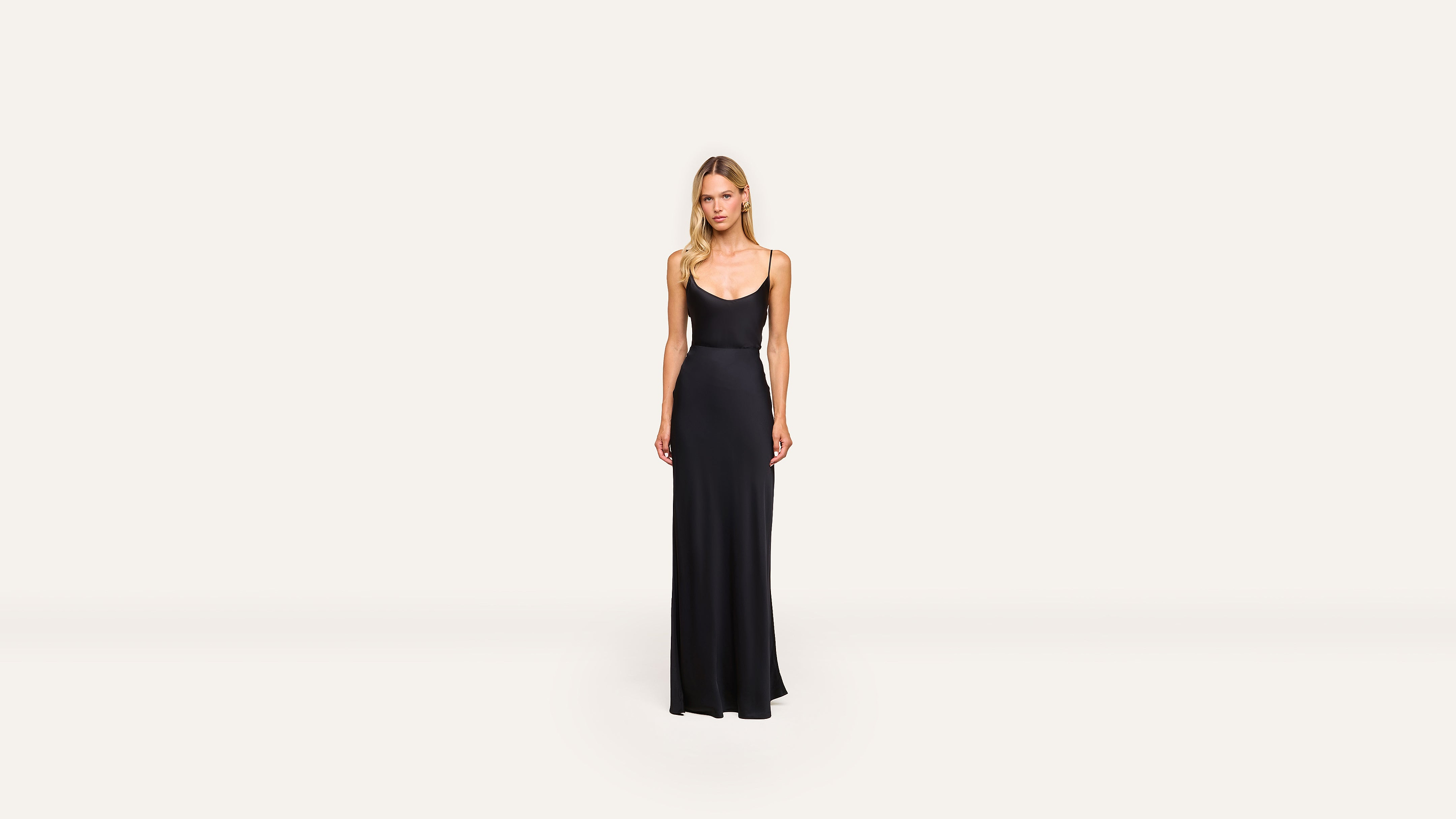 A woman stands against a plain, light background wearing a long, sleeveless black dress with a scoop neckline and minimalist style.
