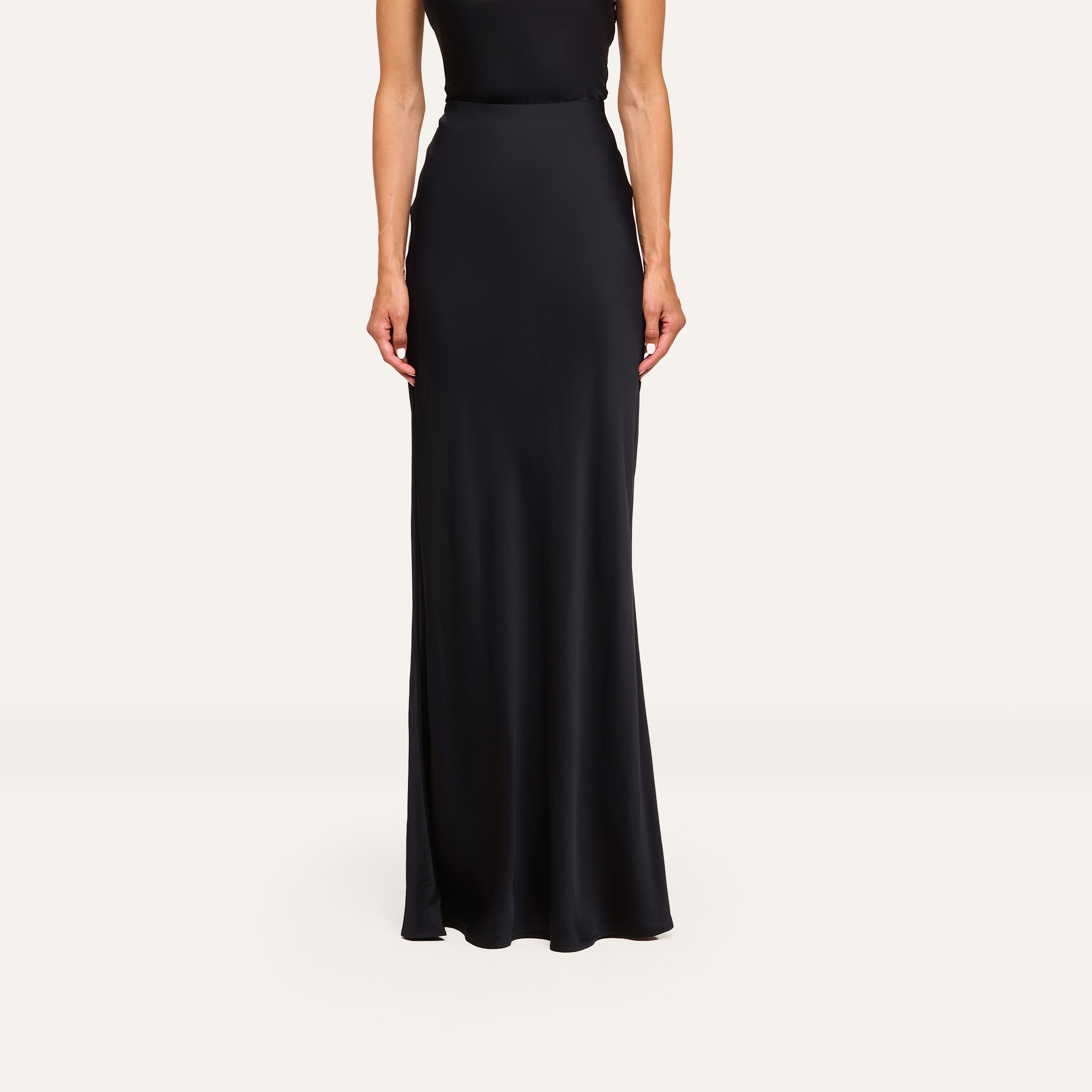 A person models the Zeta Satin Maxi Skirt—black, bias-cut, floor-length satin—standing against a plain white background with relaxed arms and head not visible.