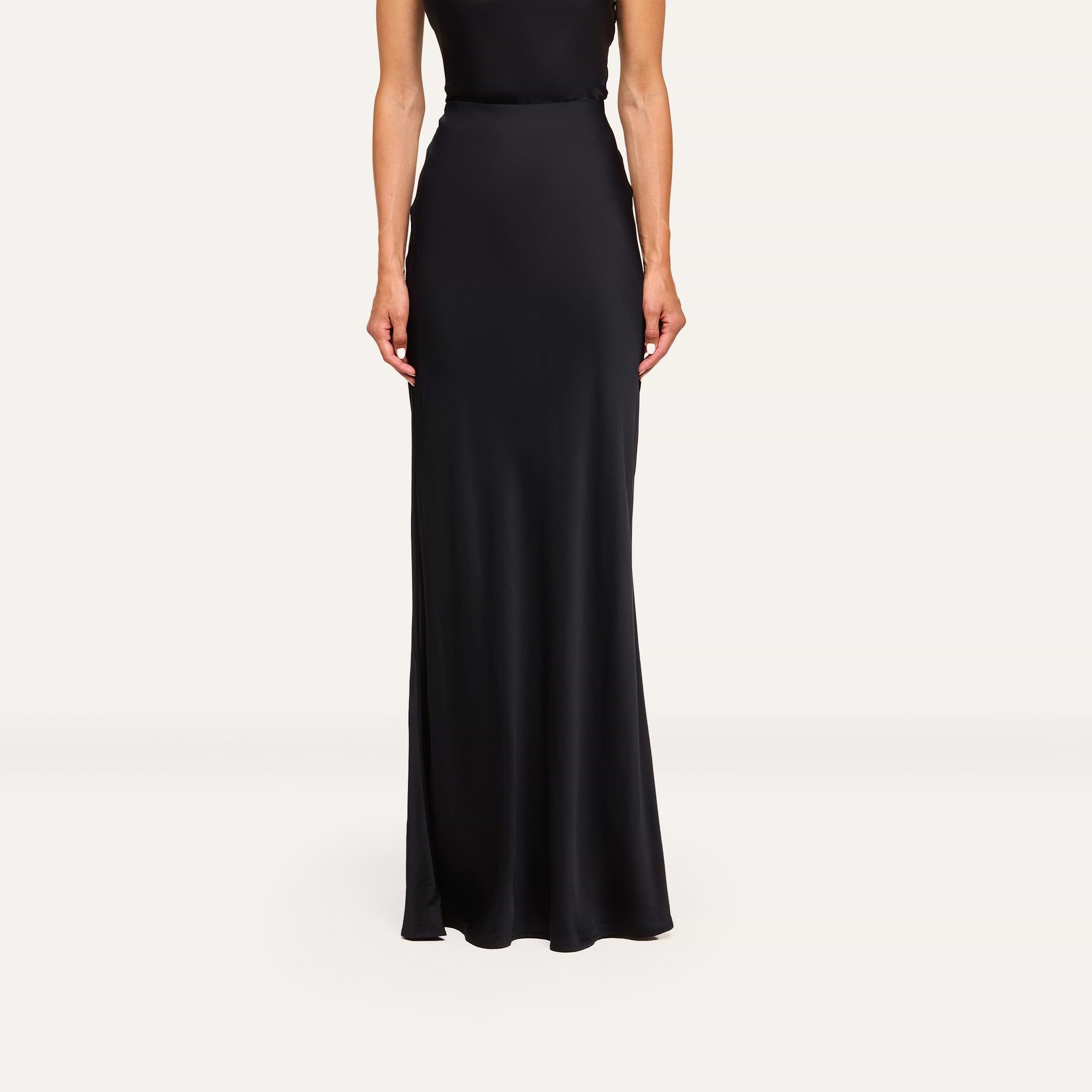 A person models the Zeta Satin Maxi Skirt—black, bias-cut, floor-length satin—standing against a plain white background with relaxed arms and head not visible.