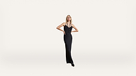 A woman in a long, fitted black dress with thin straps stands confidently with her hands on her hips against a plain white background.