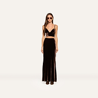 A woman wearing sunglasses stands facing forward in a dark brown velvet crop top and the Zeta Velvet Maxi Skirt with boots, against a plain light background, exemplifying a minimalist wardrobe.