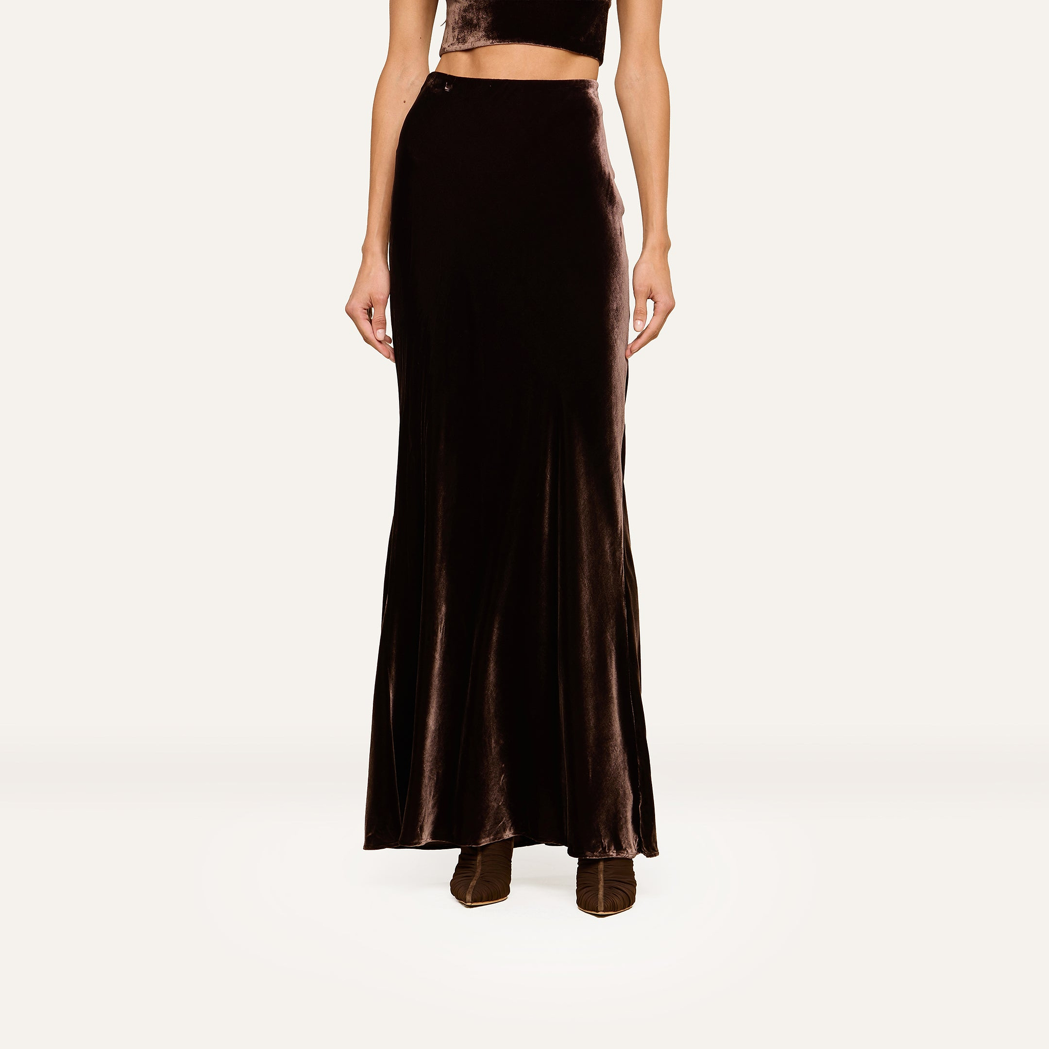 A woman wearing a sleeveless black velvet crop top and the Zeta Velvet Maxi Skirt, paired with heeled boots, stands against a plain light background.