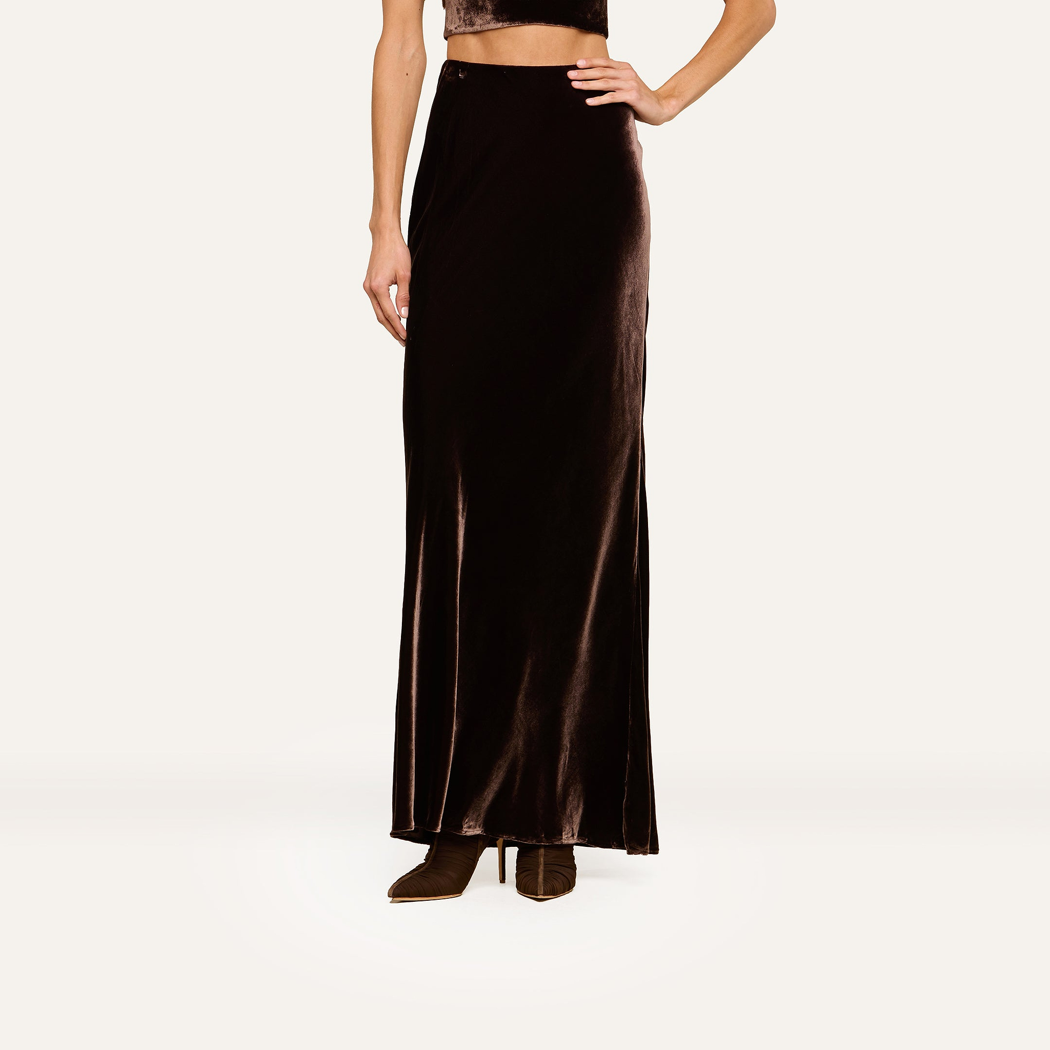 A person models the Zeta Velvet Maxi Skirt with brown boots and a cropped top, standing against a plain white background. The image focuses on the lower half of the body, highlighting a minimalist wardrobe style.