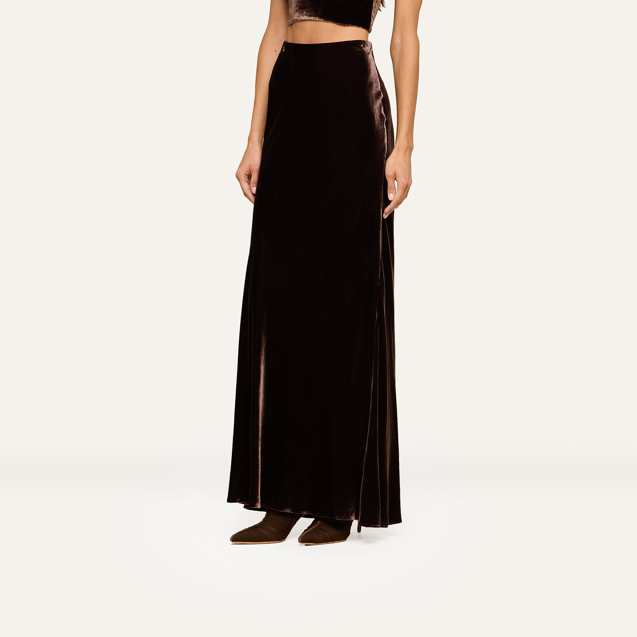 A woman models the Zeta Velvet Maxi Skirt, a black floor-length design, paired with dark shoes and a midriff-baring top against a plain white background.
