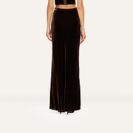 A person stands facing away, wearing the Zeta Velvet Maxi Skirt in dark brown and a black sleeveless top—an understated look that would complement a minimalist wardrobe. Only the lower half of their body is visible against a plain light background.
