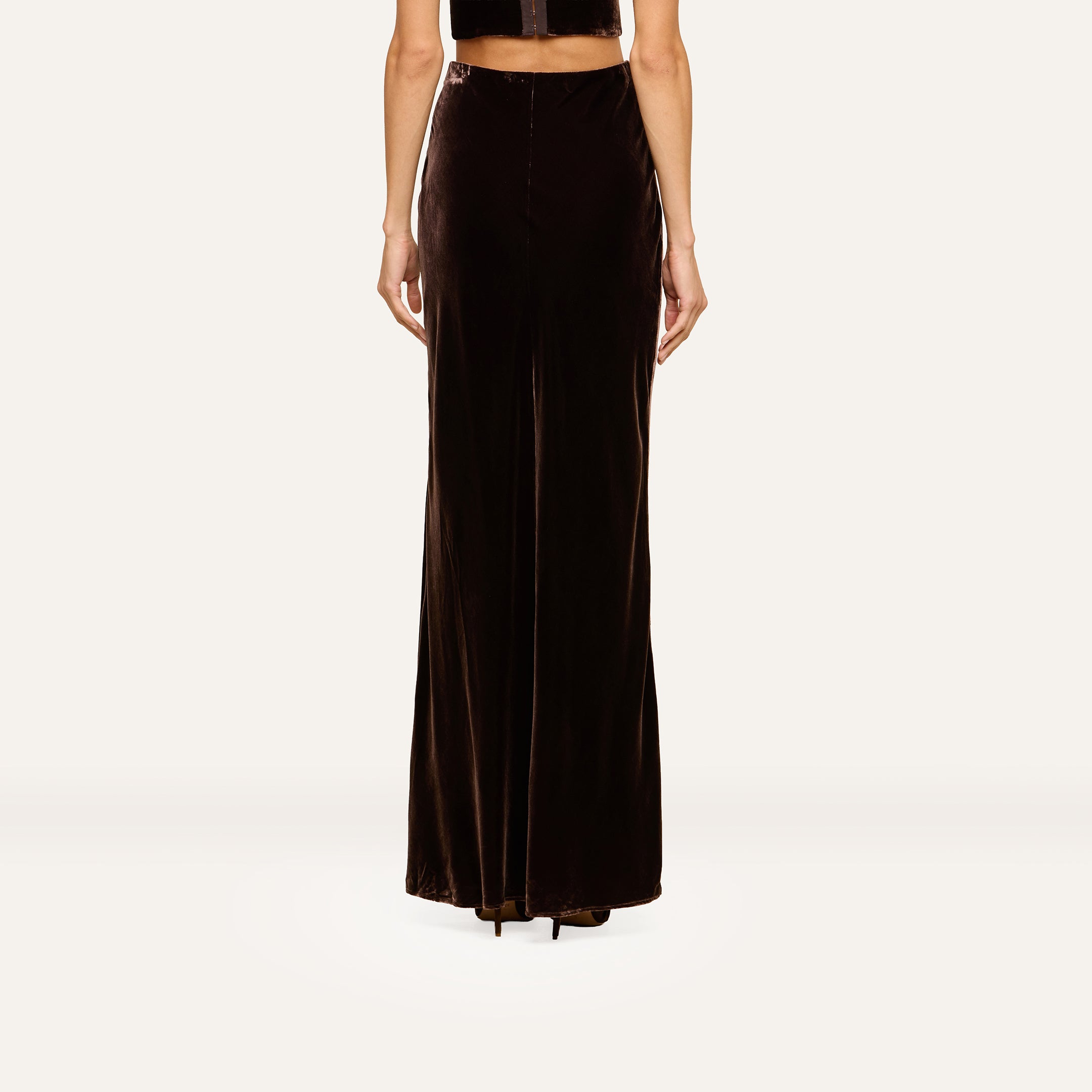 A person stands facing away, wearing the Zeta Velvet Maxi Skirt in dark brown and a black sleeveless top—an understated look that would complement a minimalist wardrobe. Only the lower half of their body is visible against a plain light background.