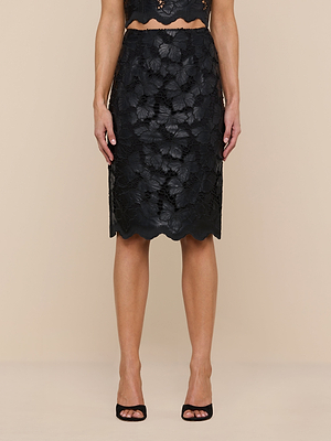 A woman stands against a plain beige background wearing the Royal Pencil Skirt, a black knee-length classic pencil skirt with a textured floral design and scalloped hem, paired with black open-toe heels.