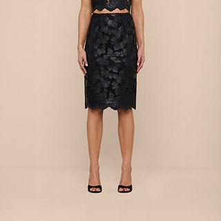 A woman models the Royal Pencil Skirt, crafted in black textured vegan leather, paired with black open-toed heels. She stands against a beige background with her arms relaxed at her sides and her head out of frame.