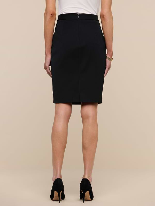 A woman stands facing away, wearing a white top and the Olyvia Pencil Skirt—a black, curve-skimming pencil skirt crafted from stretch suit fabric—paired with black high heels. The background is plain and white.