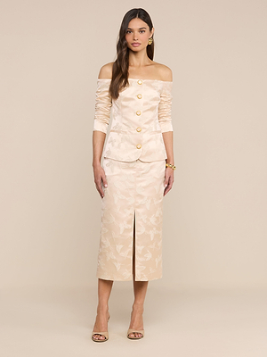 A woman stands against a neutral background wearing the Alice Blazer Top in cream jacquard satin with an off-the-shoulder neckline and ruched sleeves, paired with a matching midi skirt, heeled sandals, gold jewelry, and long hair.
