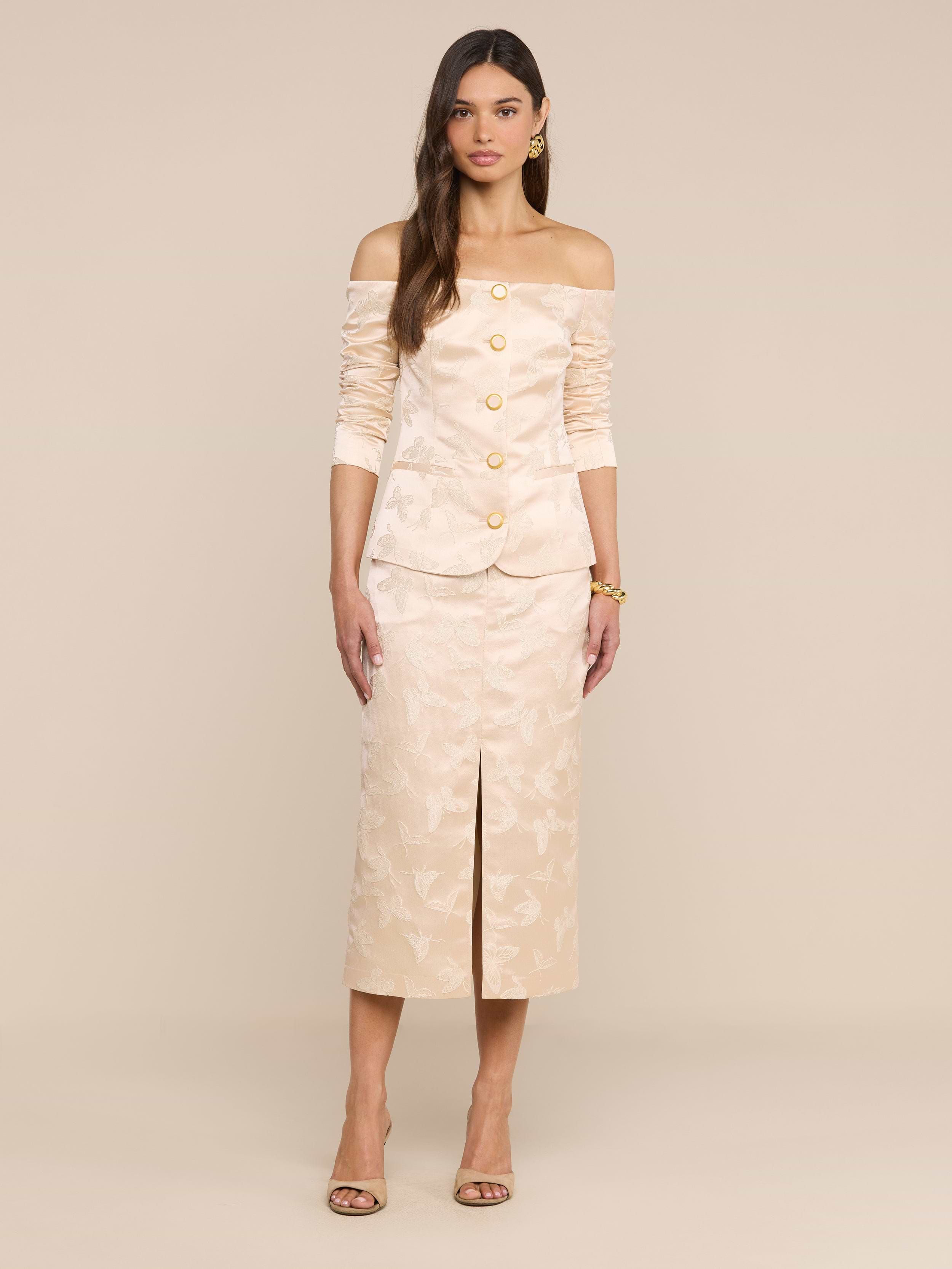 A woman stands against a plain white background wearing the Alice Blazer Top, a cream-colored, off-the-shoulder blazer with gold buttons, paired with a matching below-the-knee skirt featuring a front slit and heels.
