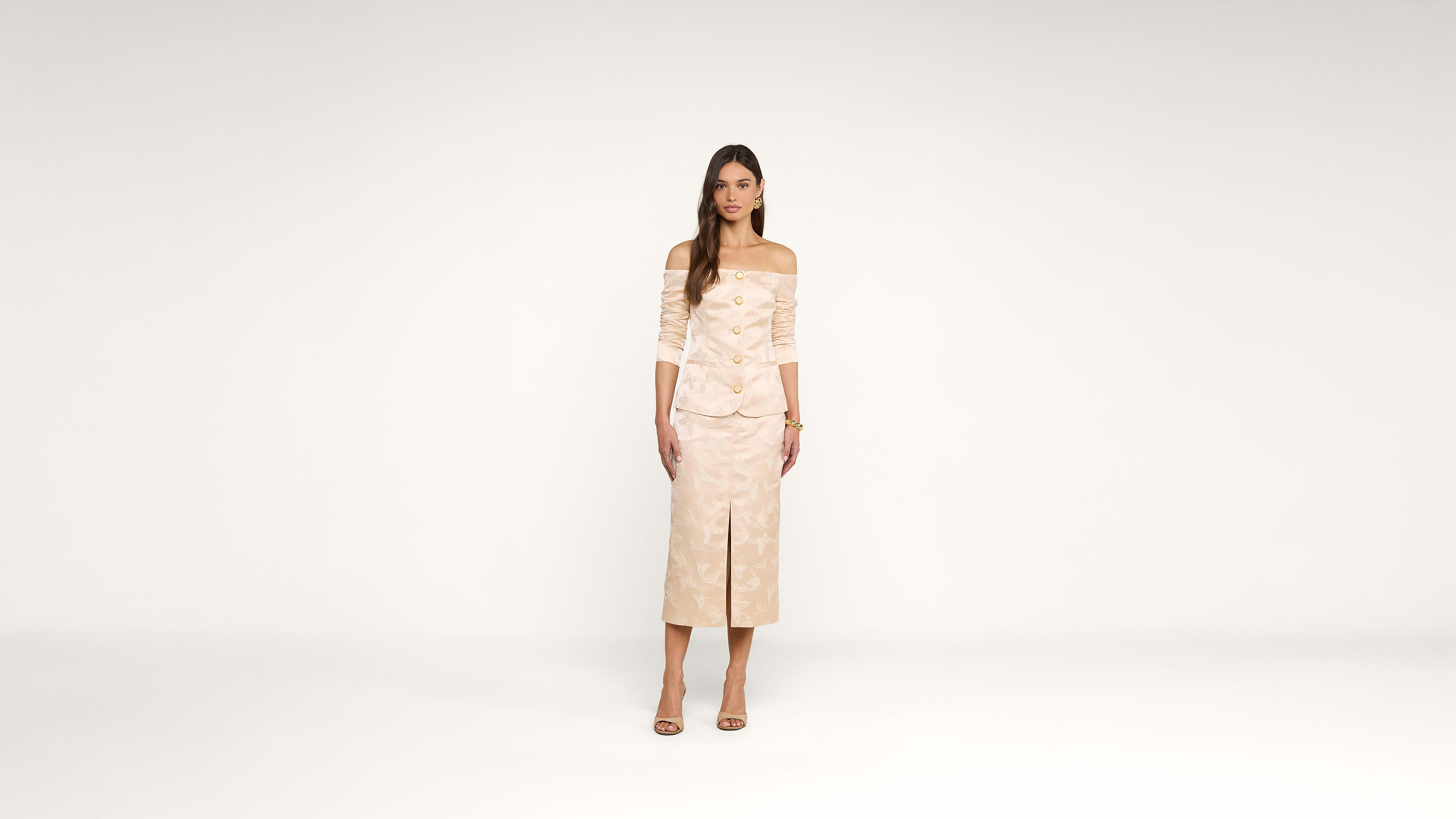 A woman in an off-shoulder beige suit with gold buttons and a matching skirt stands facing forward in a plain white studio. She is wearing beige heels and has long straight hair.