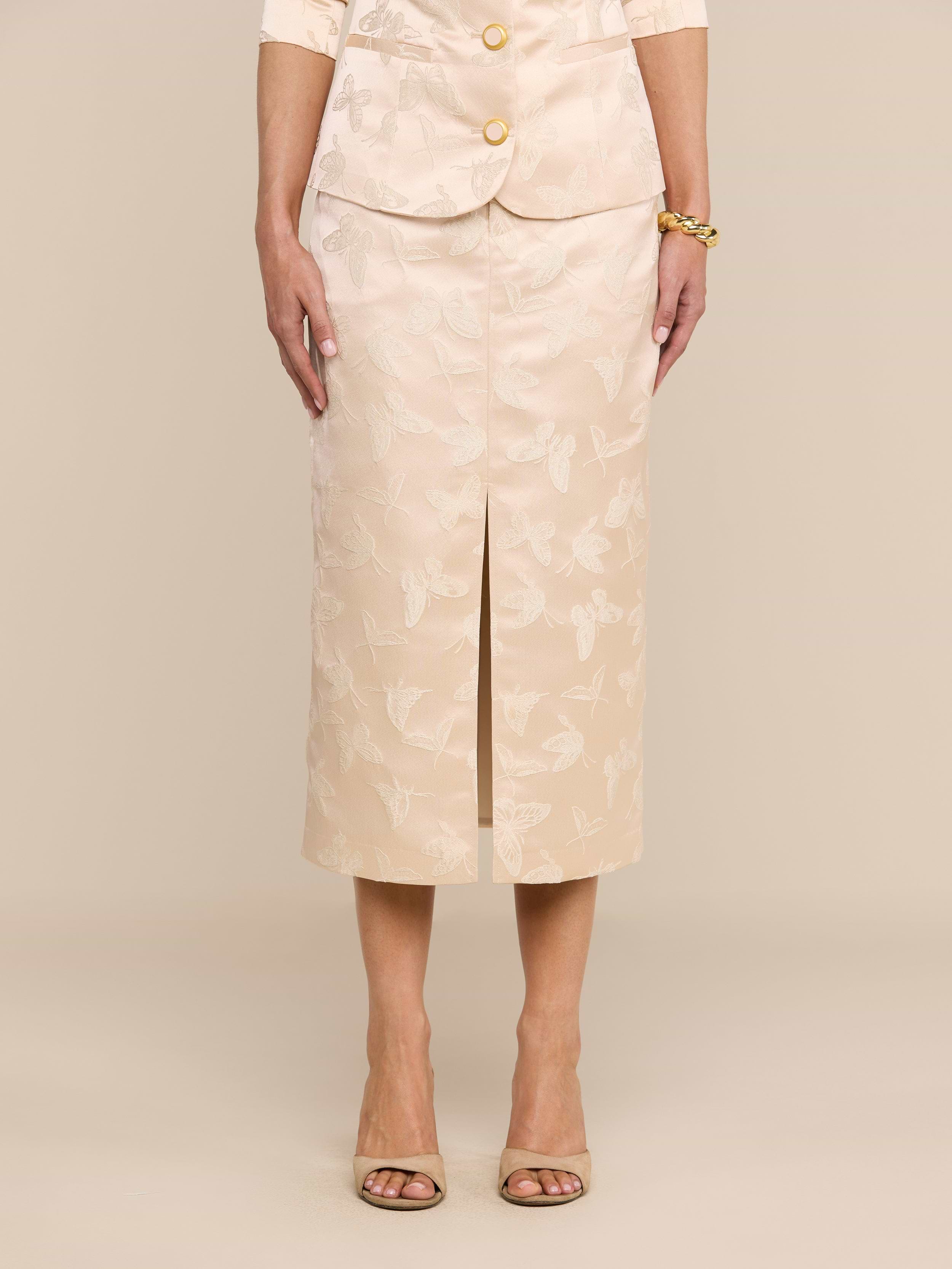 A woman stands facing forward against a plain white background, wearing the Madonna Midi Skirt with a beige butterfly jacquard floral pattern as part of a matching skirt suit. Her hands rest relaxed at her sides, and she accessorizes with open-toed heels and a bracelet.