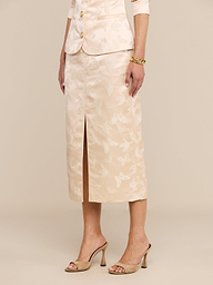 A woman stands against a plain white background, arms at her sides, wearing the Madonna Midi Skirt—a light beige skirt in butterfly jacquard with a floral pattern and a front slit—paired with high-heeled sandals.