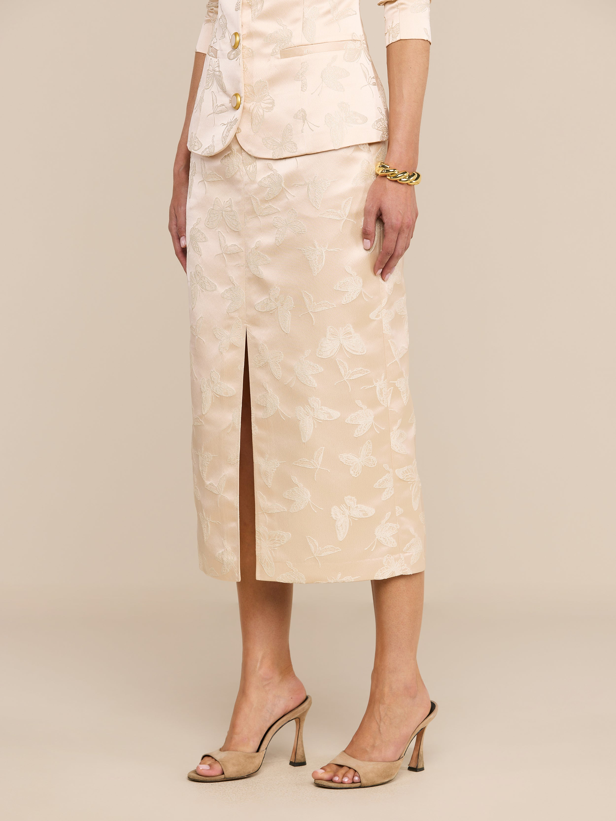 A woman stands against a plain white background, arms at her sides, wearing the Madonna Midi Skirt—a light beige skirt in butterfly jacquard with a floral pattern and a front slit—paired with high-heeled sandals.