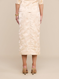 A woman stands facing away, wearing the Madonna Midi Skirt in light beige with a matching top featuring elbow-length sleeves. She pairs the outfit with high-heeled shoes against a plain white background.