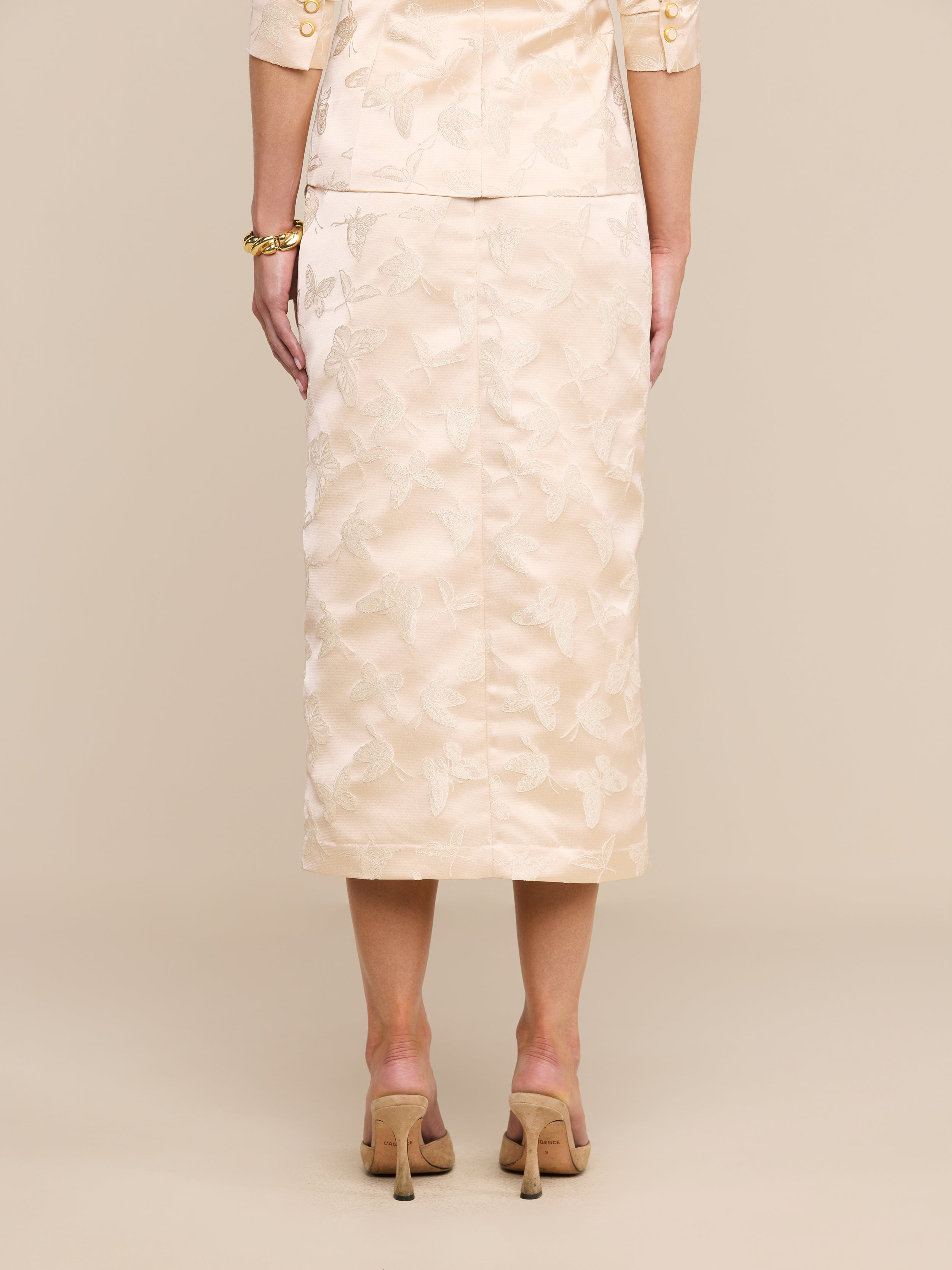 A woman stands facing away, wearing the Madonna Midi Skirt in light beige with a matching top featuring elbow-length sleeves. She pairs the outfit with high-heeled shoes against a plain white background.