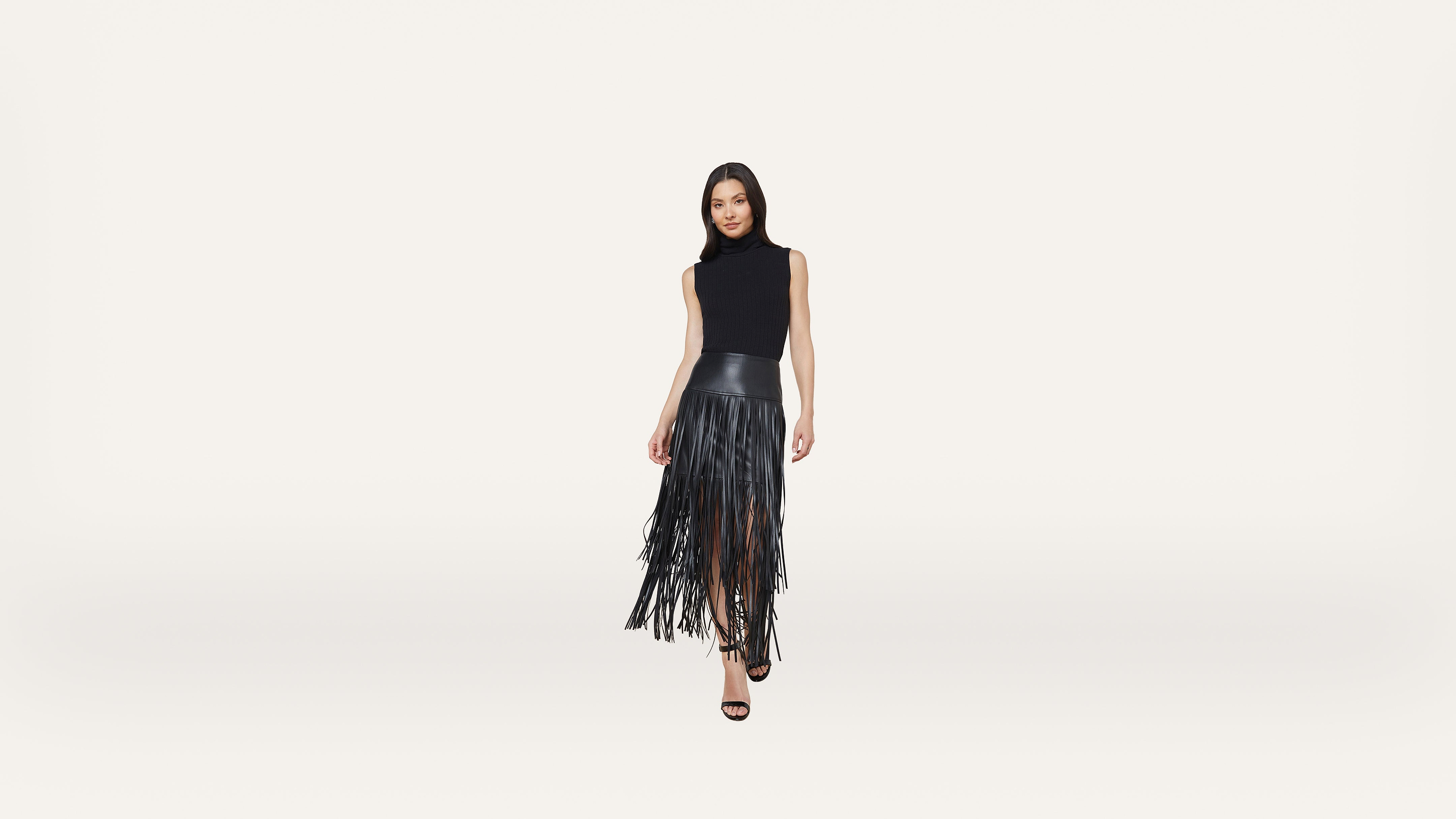 Woman standing and posing in a sleeveless black top and a black fringe skirt with high heels, against a plain white background.