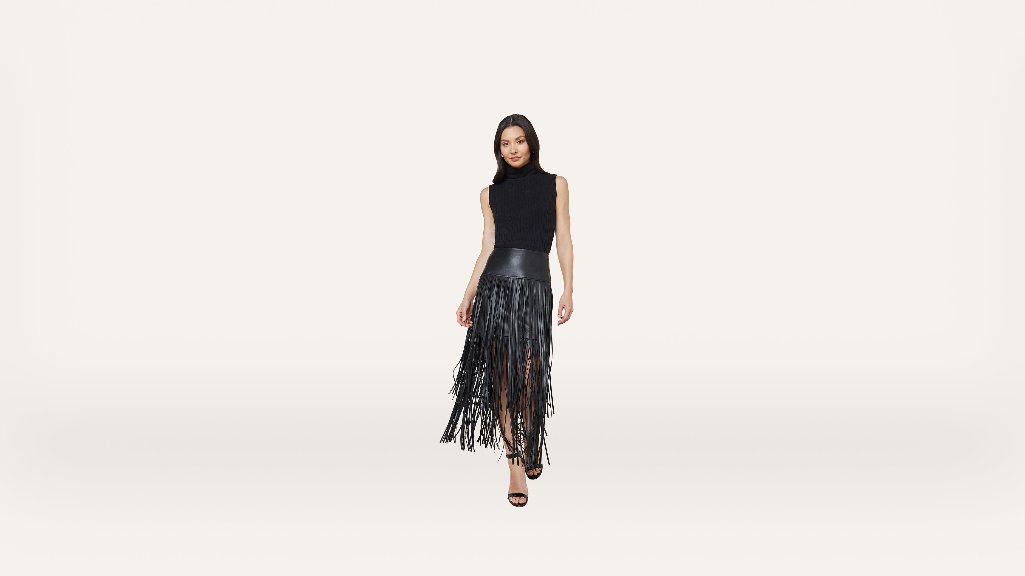 Woman standing and posing in a sleeveless black top and a black fringe skirt with high heels, against a plain white background.