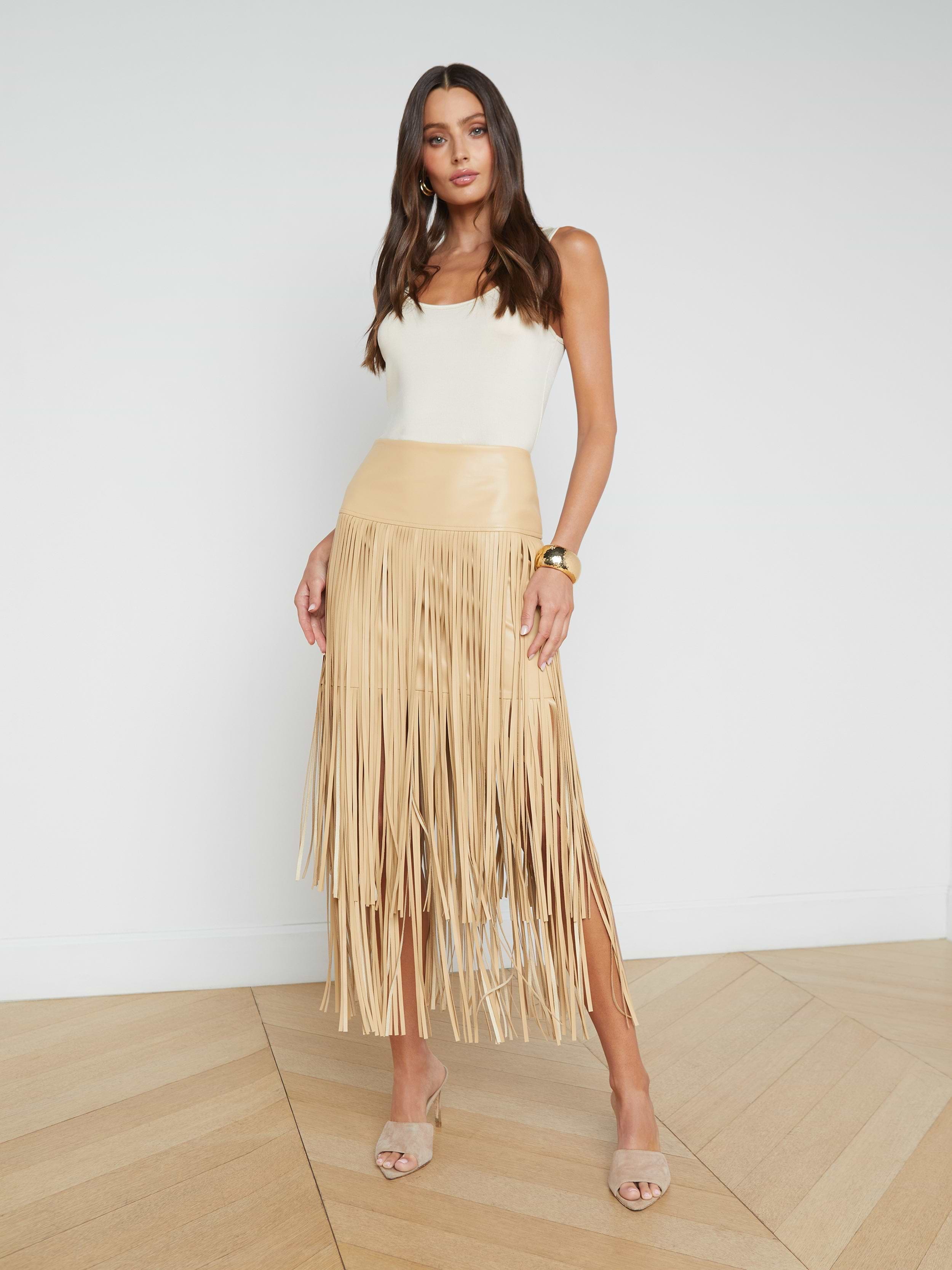 A woman stands facing forward in high heels against a plain white background, wearing a white sleeveless top and the tan Karolina Fringe Skirt made from vegan leather.