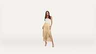 A woman stands against a white background, wearing a white sleeveless top and a tan fringed skirt with open-toe heels. She has long hair and is looking at the camera with a relaxed pose.