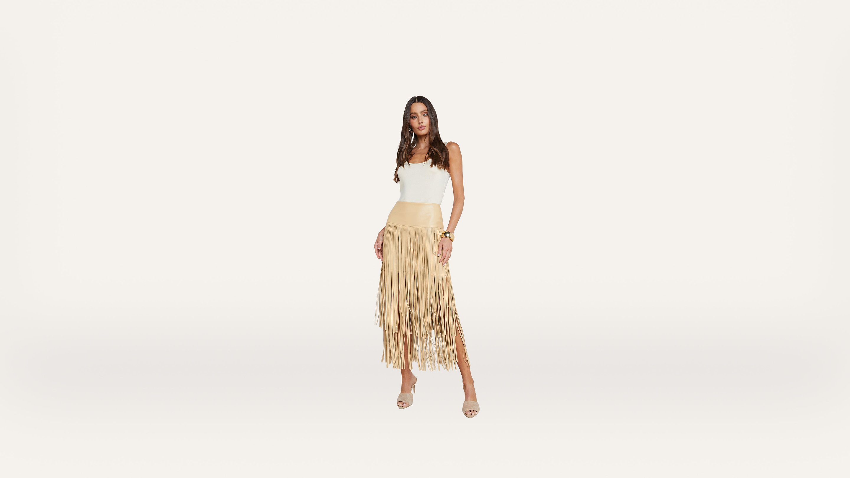 A woman stands against a white background, wearing a white sleeveless top and a tan fringed skirt with open-toe heels. She has long hair and is looking at the camera with a relaxed pose.