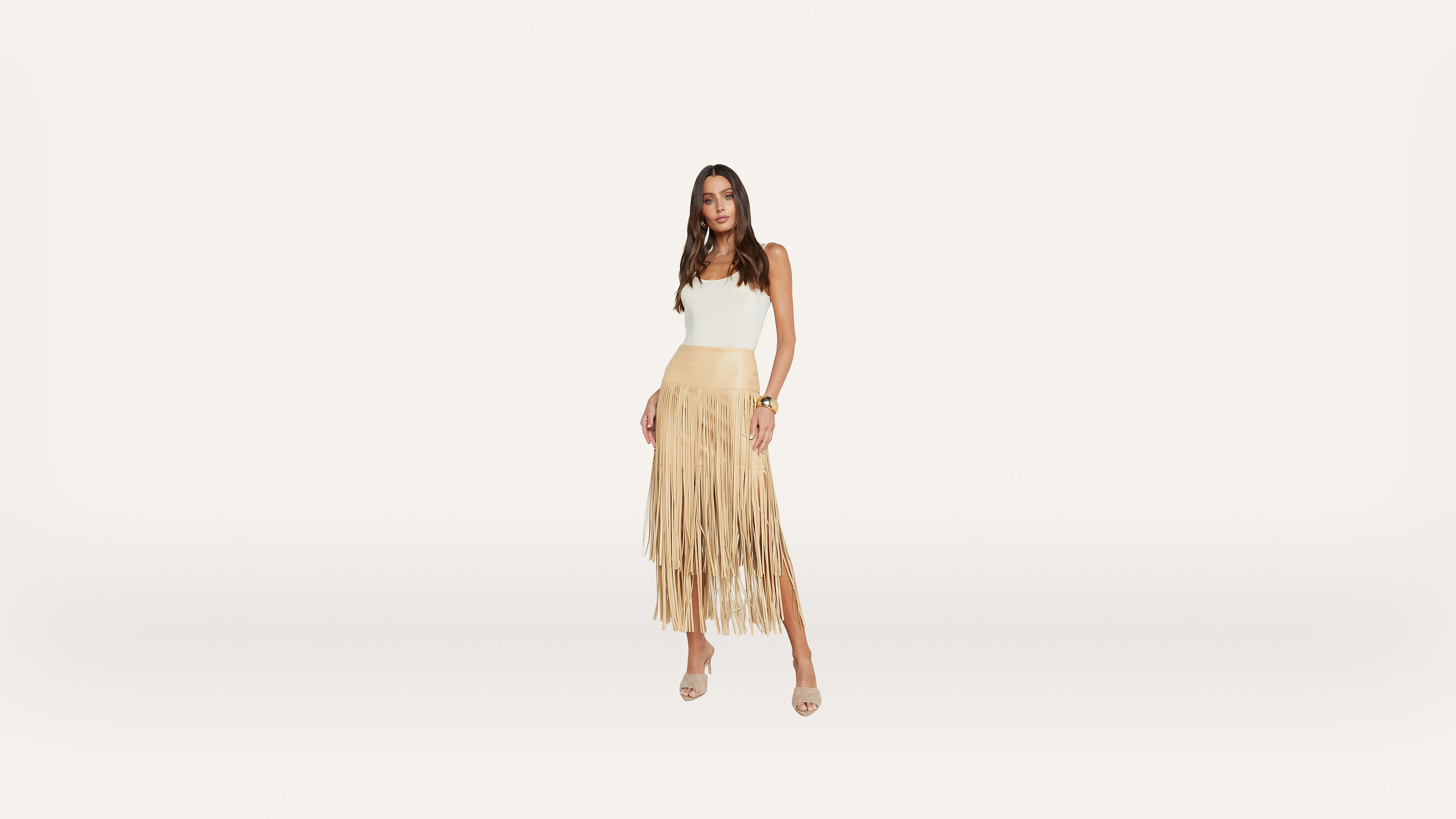 A woman stands against a white background, wearing a white sleeveless top and a tan fringed skirt with open-toe heels. She has long hair and is looking at the camera with a relaxed pose.