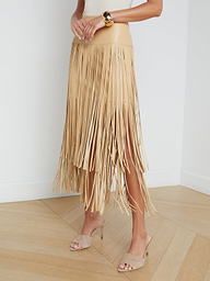 A person wearing the Karolina Fringe Skirt, a white top, beige heeled sandals, and a gold bracelet stands against a plain white background.